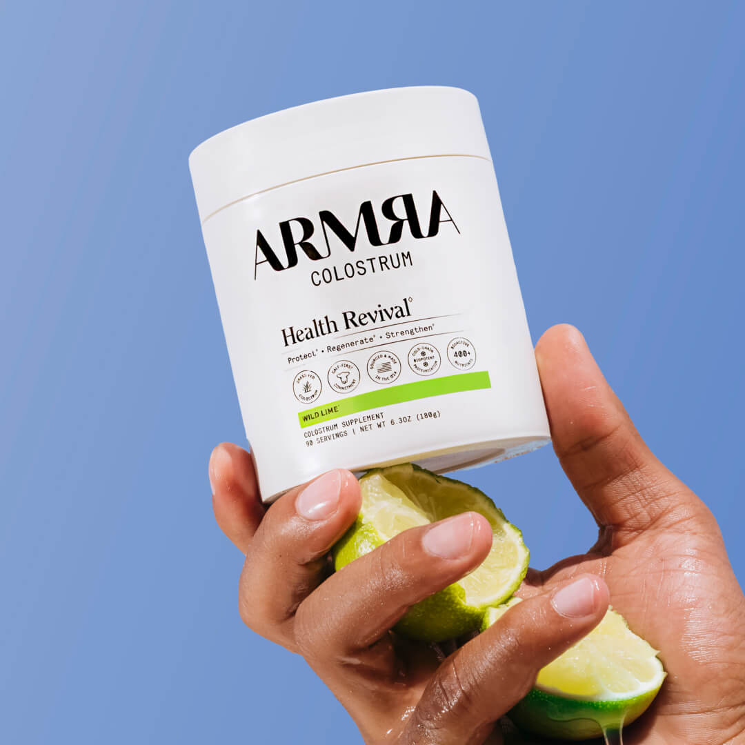 Hand holding a container of ARMRA Colostrum with limes on a blue background