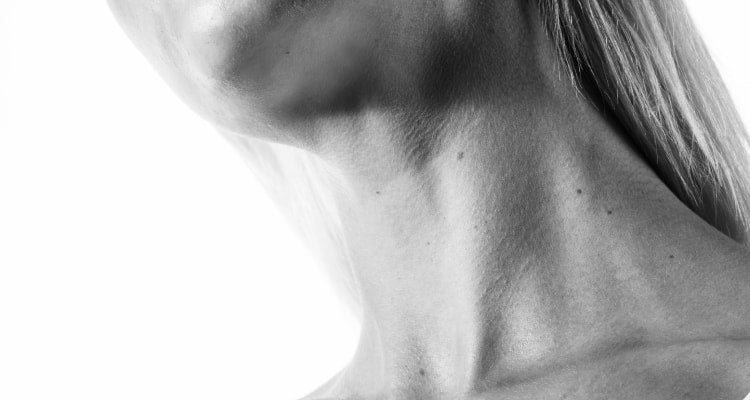 A black and white cropped image showing a womans chest, neck, chin tilted up, and lips wearing a white tank top on a white background